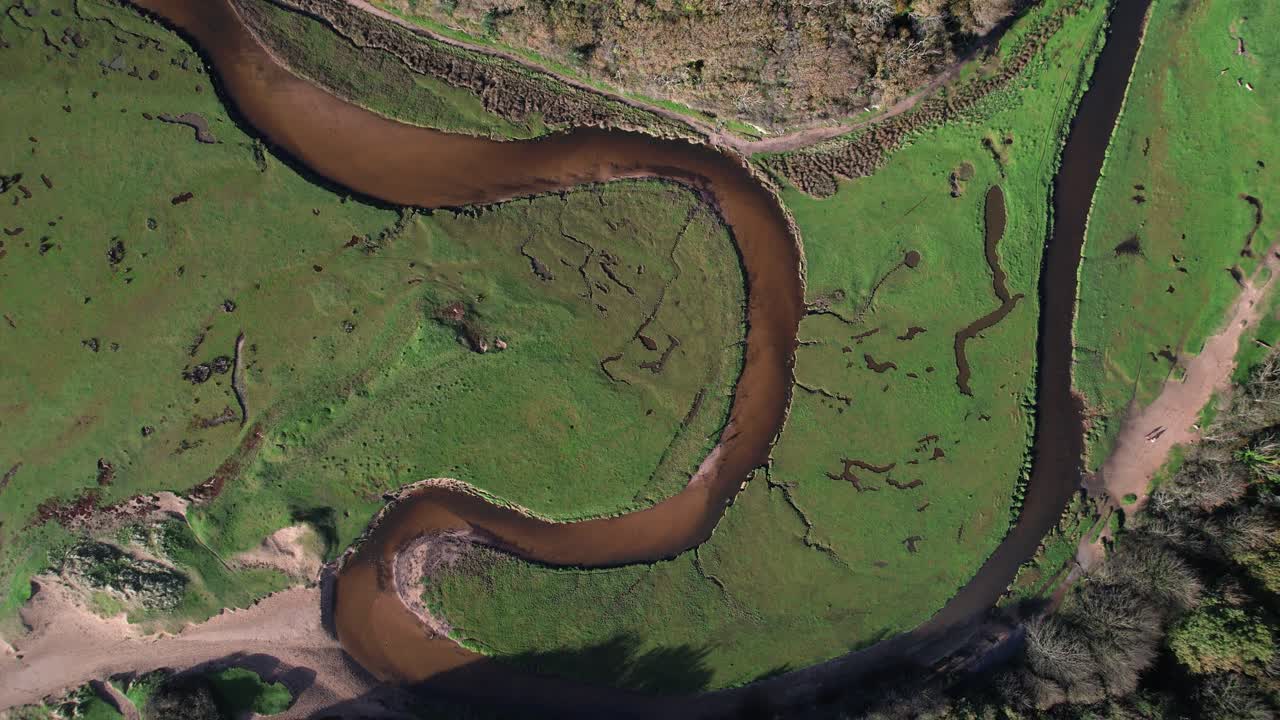Top down drone ascent revealing the winding Pennard Pill river through lush green fields near Three Cliffs Bay in Wales