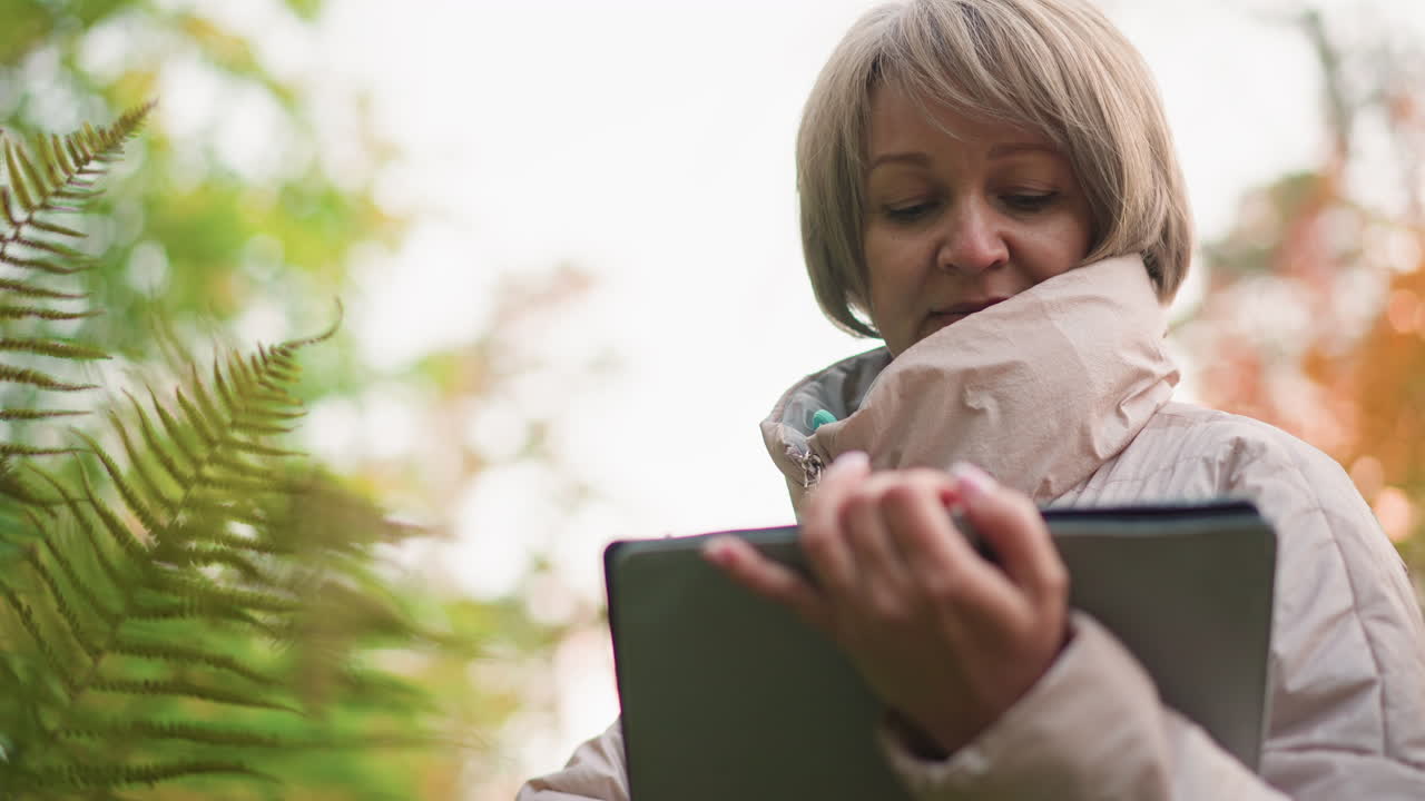 close up of female researcher observing fern leaf while recording notes on tablet in autumn forest, focused expression on face as she documents data during outdoor field study