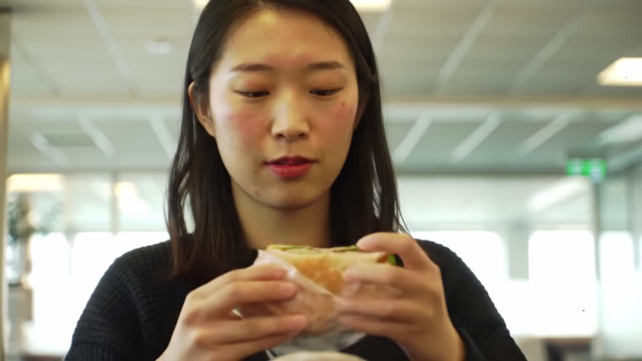 A young woman carefully unwraps and examines her sandwich in a welcoming cafe setting. It’s a busy lunch hour, filled with joyful conversations and delicious aromas.