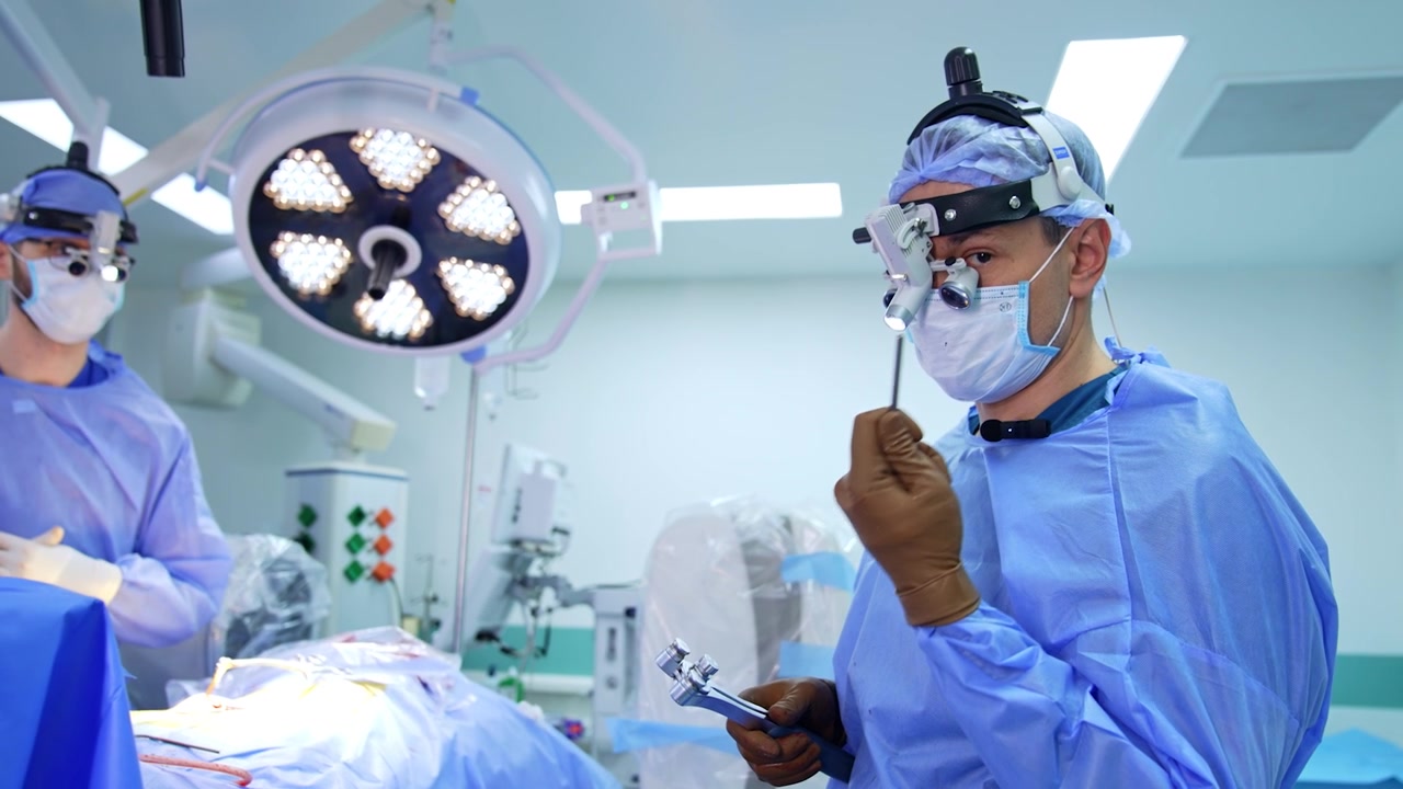 Male doctor in uniform and surgical loupes holds a tool showing it to camera. Surgeon explain usage of the instrument at neurosurgery.