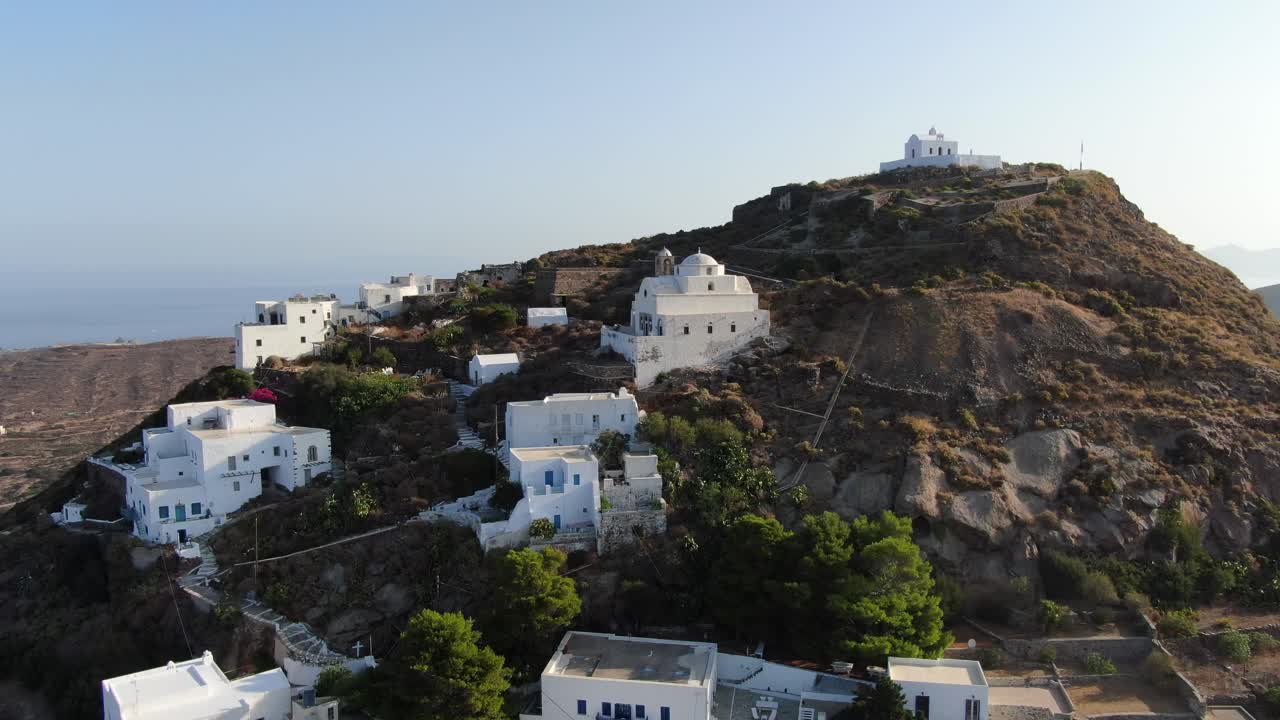 vista de drone en grecia volando frente a la ciudad griega con casas blancas en una colina marrón con una iglesia en la parte superior y el mar en el horizonte en un día soleado