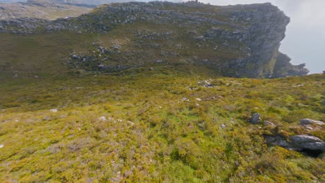 Toma-Aérea-Volando-Sobre-Un-Hermoso-Terreno-Accidentado-En-La-Montaña-De-La-Mesa-En-Sudáfrica,-Con-Ciudad-Del-Cabo-Visible-En-La-Distancia-Debajo