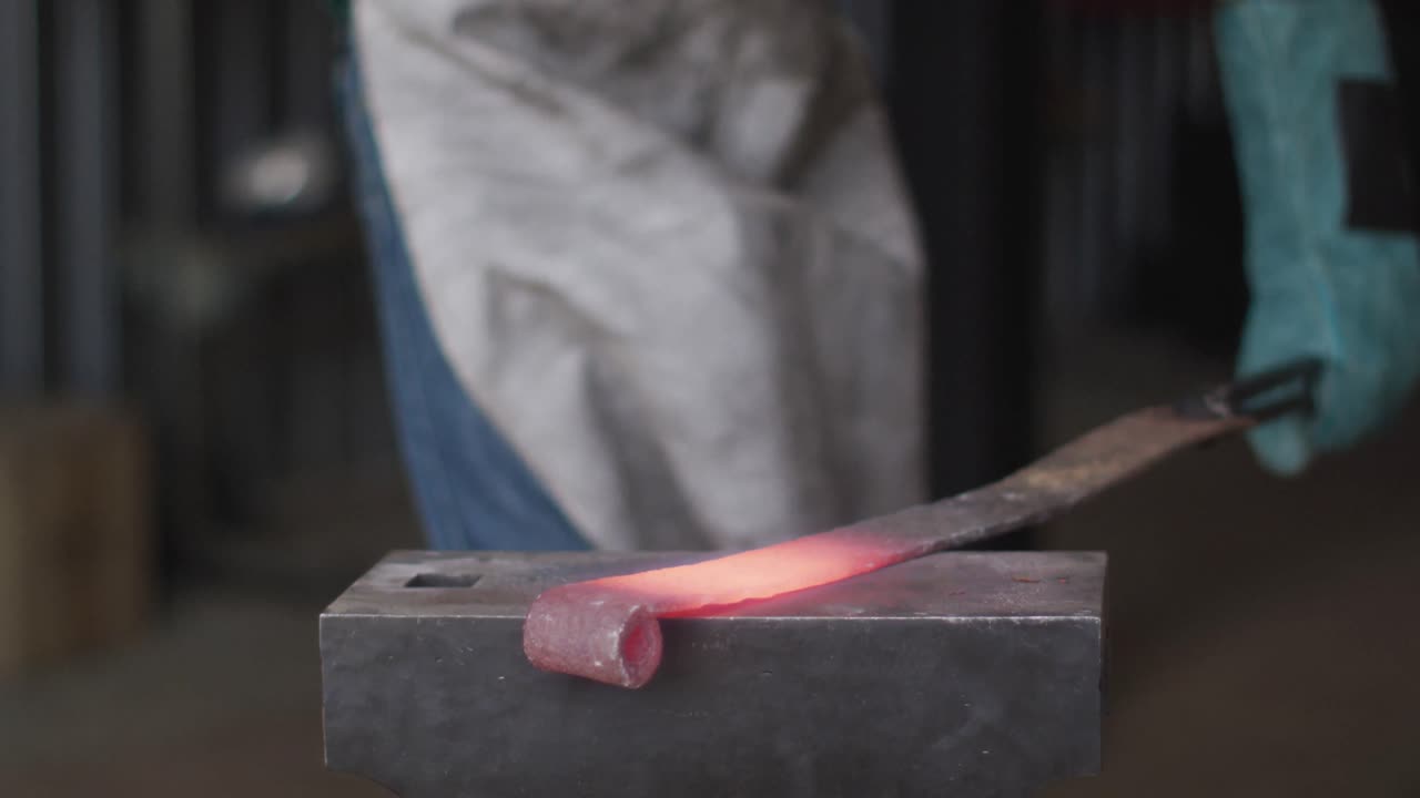 Midsection of caucasian male blacksmith wearing safety glasses, hammering hot metal tool on anvil