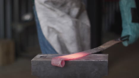 Midsection-of-caucasian-male-blacksmith-wearing-safety-glasses,-hammering-hot-metal-tool-on-anvil