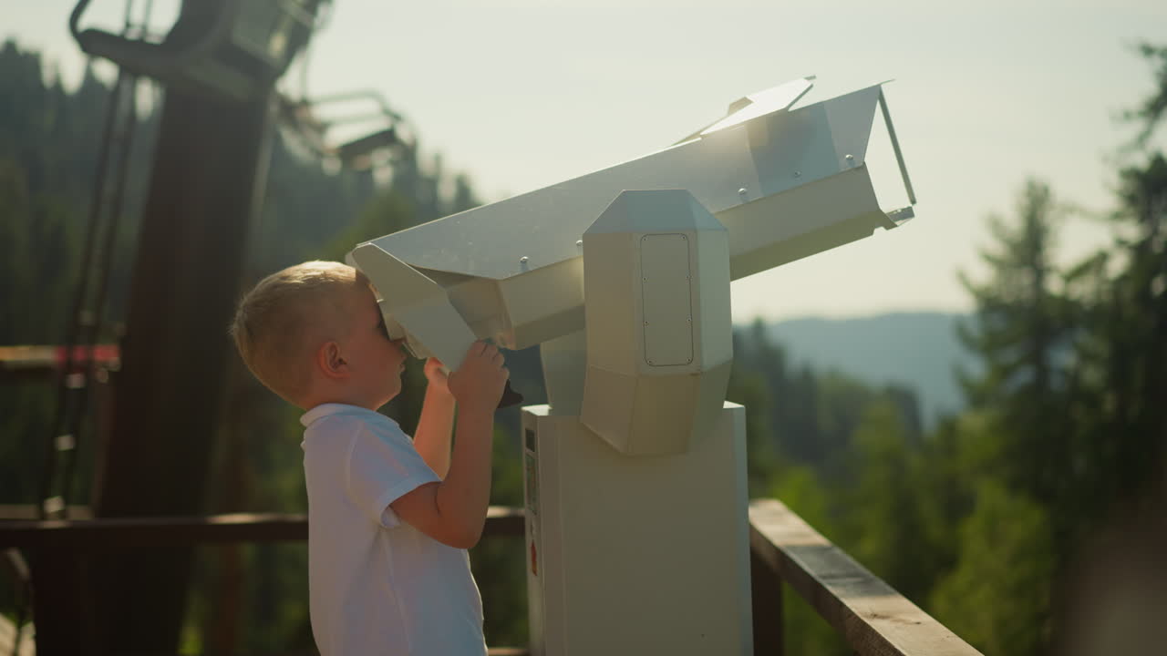 Focused toddler boy looks at sky through huge binoculars for tourists against moving cable cars. Relaxation in nature and admiration of beauty slow motion