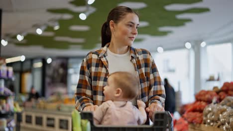 Close-up-a-confident-brunette-girl-in-a-plaid-shirt-carries-her-little-daughter-on-a-cart-while-shopping-and-inspecting-goods-in-a-supermarket