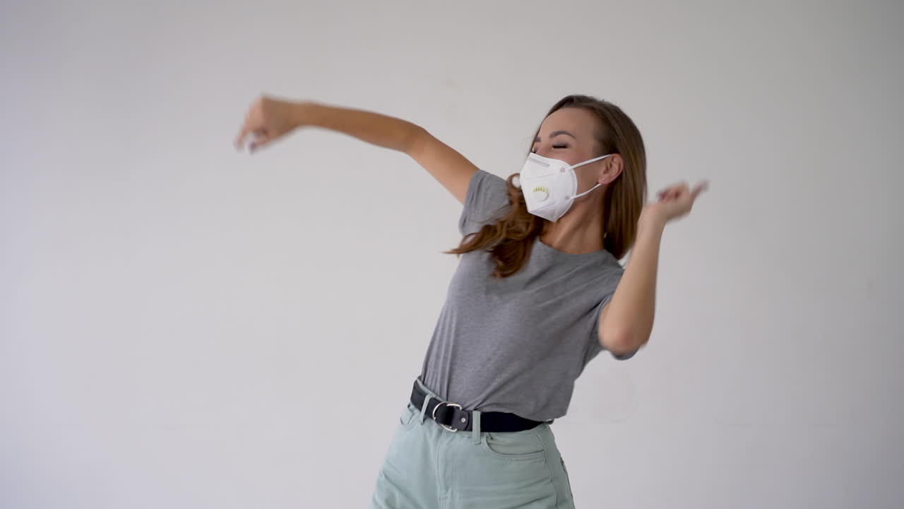 ⁇ 색 배경에 의료 마스크를 착용하고 춤을 추는 즐거운 젊은 여성. 코로나바이러스 격리 조치의 끝.