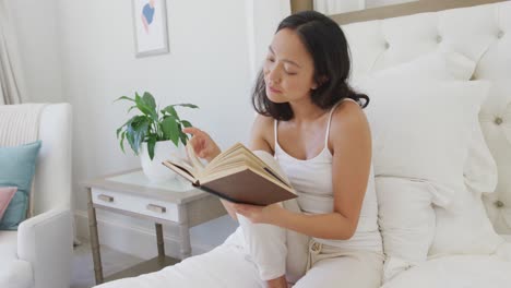 happy asian woman sitting on bed and reading book, in slow motion