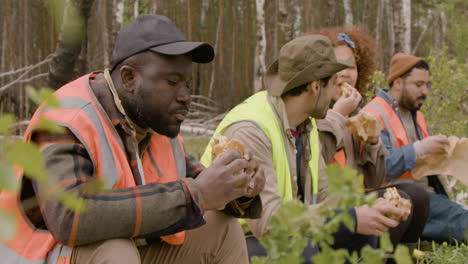 Side-view-of-a-group-of-multiethnic-ecologist-activists-eating-and-talking-in-a-break-sitting-on-a-trunk-in-the-forest