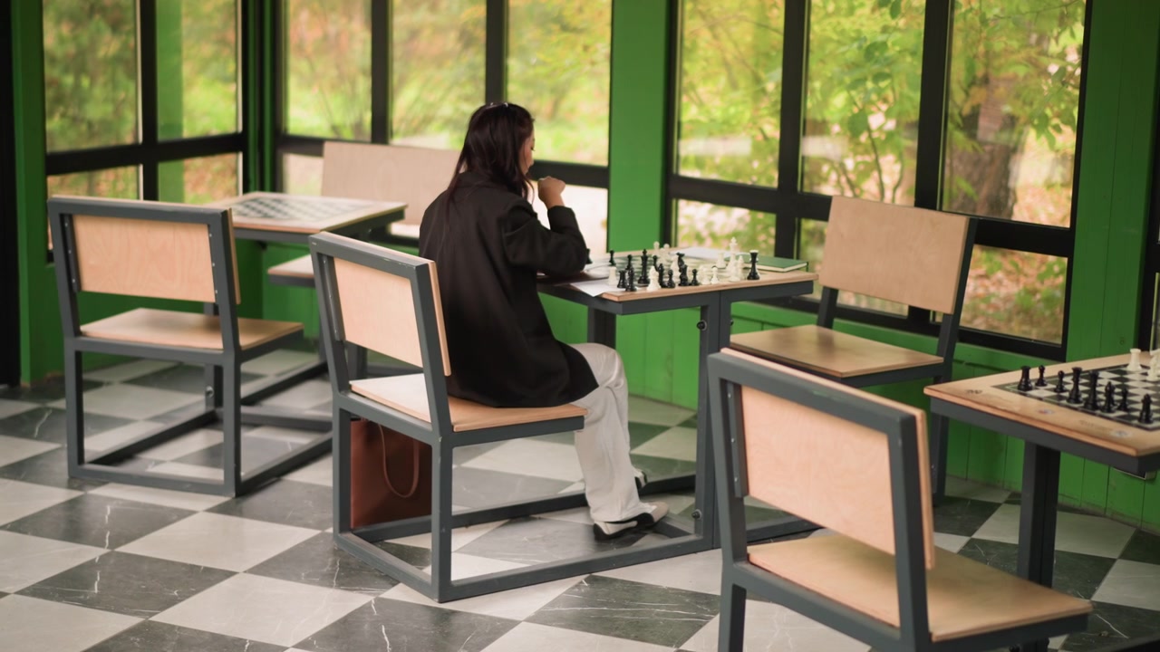 Young woman plays chess at table in indoor setting with large windows showing greenery outside. Woman is seated on metal and wood chair wearing dark clothing while interacting with game