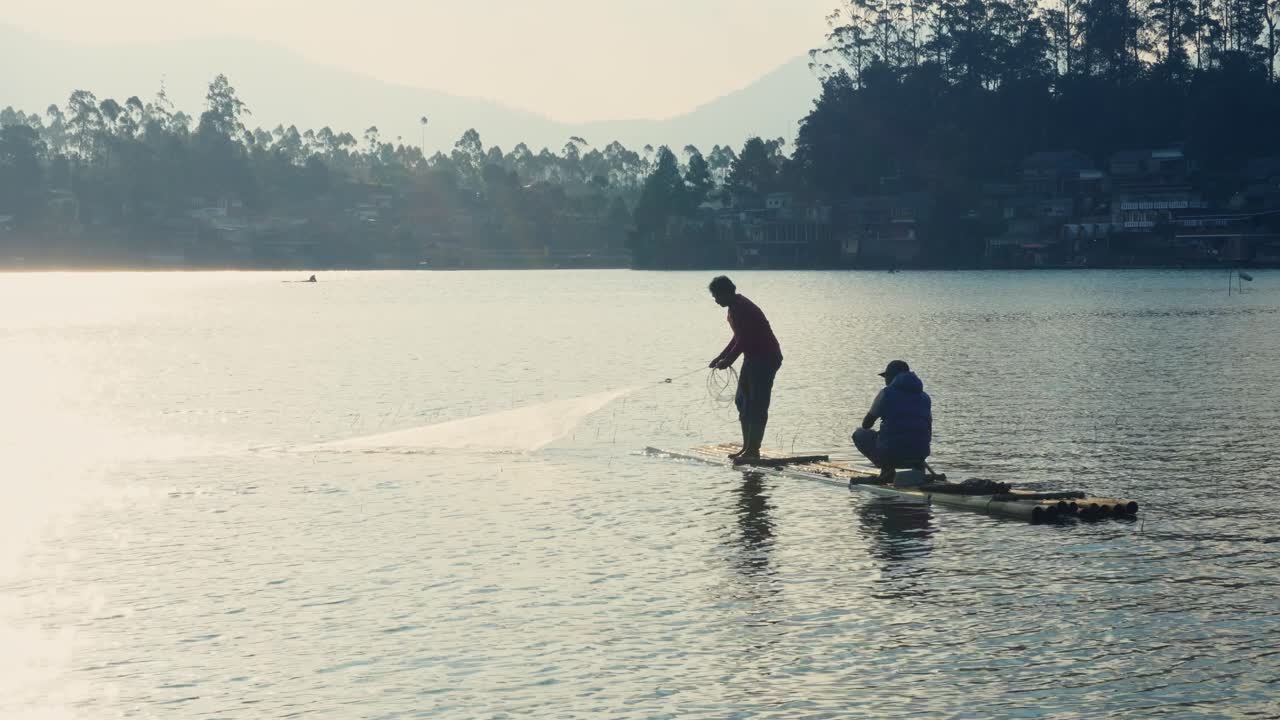Two Fishermen On Bamboo Raft In Wide Lake Under Bright Sky Throwing Fishing Net