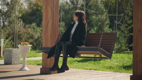 lady in black coat and leather boots sitting on wooden swing chair in a public park, enjoying bright sunlight, with green park scenery and blurred background of trees and flowers