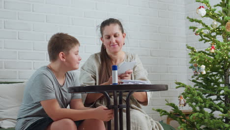 mother carving paper with scissors on table while son in gray shirt sits nearby staring attentively, cozy indoor scene featuring decorated christmas tree with ornaments in background