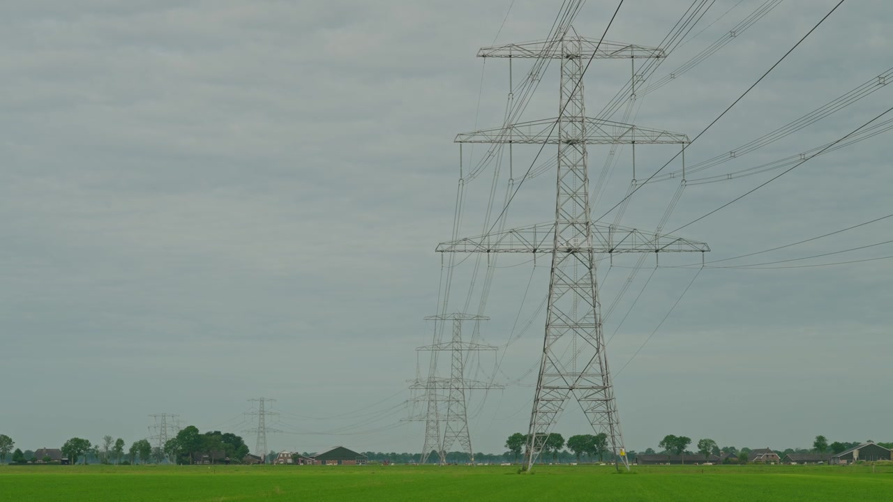 Static shot of tall electrical pylons lined across flat agricultural farmland in the Netherlands. Vibrant green grass contrasts with a moody grey overcast sky in a wide rural landscape.