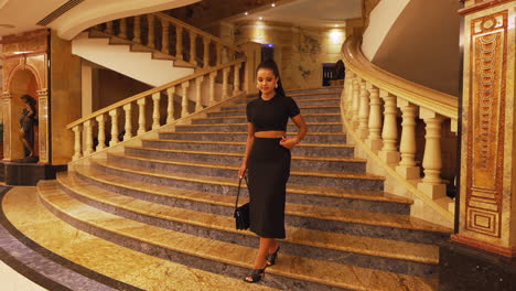 joven y encantadora modelo de dama morena caminando por lujosas escaleras interiores y dando la vuelta, vestida de negro