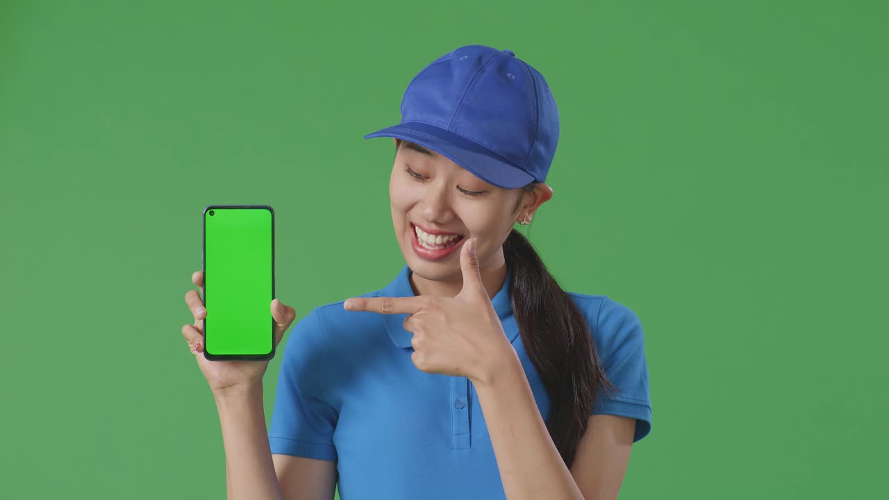primer plano de una mensajera asiática en uniforme azul sonriendo y señalando a un teléfono inteligente de pantalla verde mientras está de pie en el fondo de la pantalla verde en el estudio
