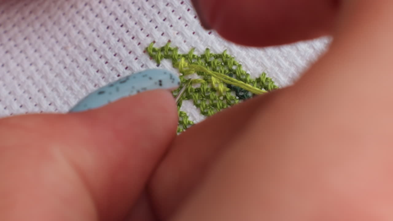 능숙한 장인이 초록색 실을 꿰어 십자수 원단을 통과시키는 극단적인 클로즈업, 깔끔한 X자 패턴과 질감을 보여주는 정확한 손놀림, 느리고 체계적인 움직임