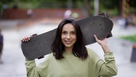 a young brunette girl stands in the background of a park with a skateboard and looks into the camera. portrait of a girl