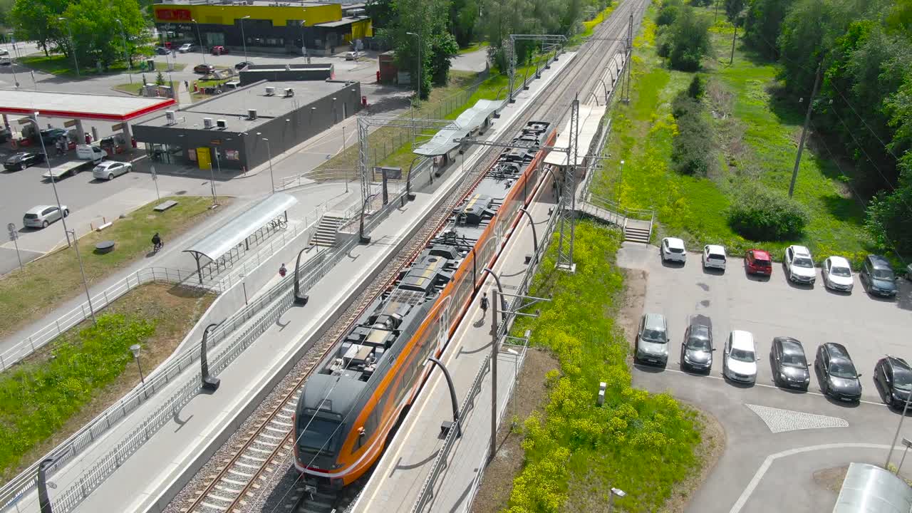 Moving aerial drone footage of an orange Estonian train arriving in Laagri train station, people boarding and getting off and train continues to move with drone moving along with it. Top view of train