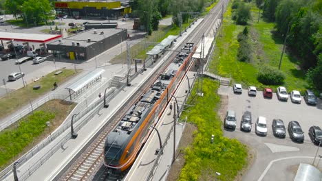 moving aerial drone footage of an orange estonian train arriving in laagri train station, people boarding and getting off and train continues to move with drone moving along with it. top view of train