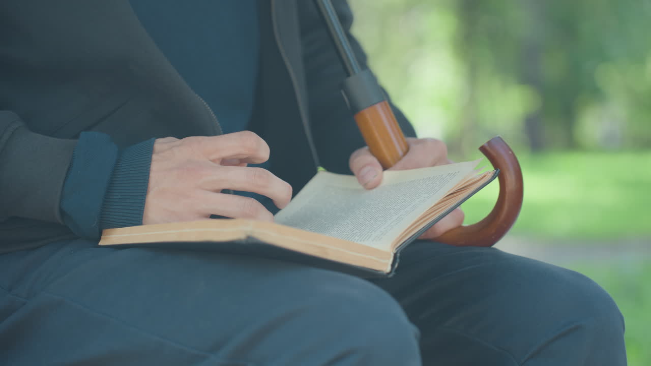 Yaşlı bir adam bankta kitap okuyor, elleri yıpranmış sayfaları çeviriyor, bastonu dizinin yanına asılı, ağaçların arasından süzülen yumuşak güneş ışığı, ellerin ve ciltsiz kitabın yakın çekimi, dingin park atmosferi, dokulu kazak, yavaş