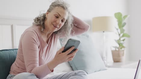 Glückliche-ältere-Kaukasische-Frau,-Die-Auf-Dem-Bett-Sitzt-Und-Auf-Dem-Smartphone-Spricht,-Zeitlupe