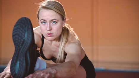 portrait of a focused blonde sportswoman warming up and stretching legs in an indoor sport facility