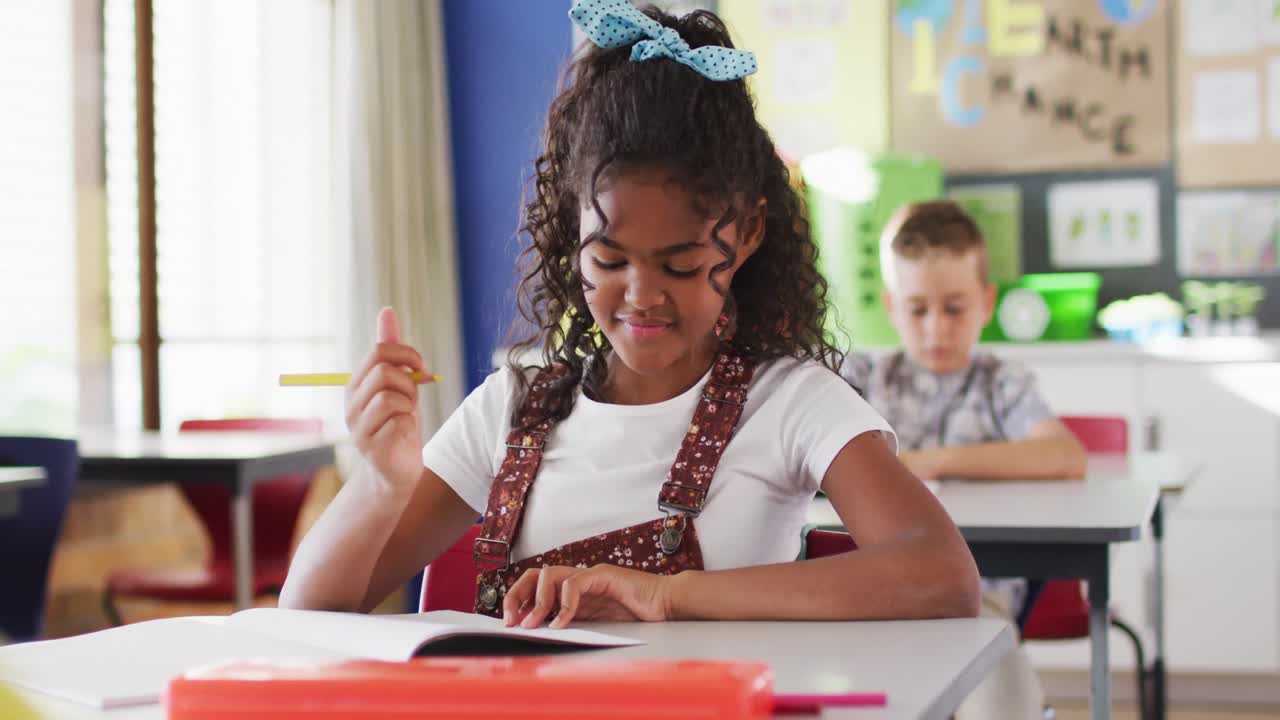 Portrait of happy mixed race schoolgirl sitting at classroom, making notes, looking at camera