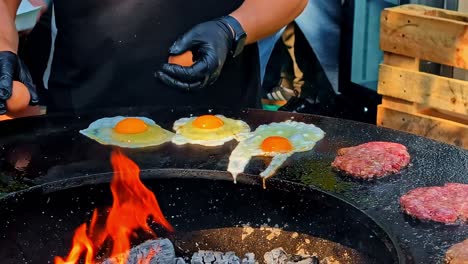 a street food vendor cooks fried eggs and burger patties on a large, round fire pit grill at an outdoor market in riga, latvia, showcasing a popular festival food experience