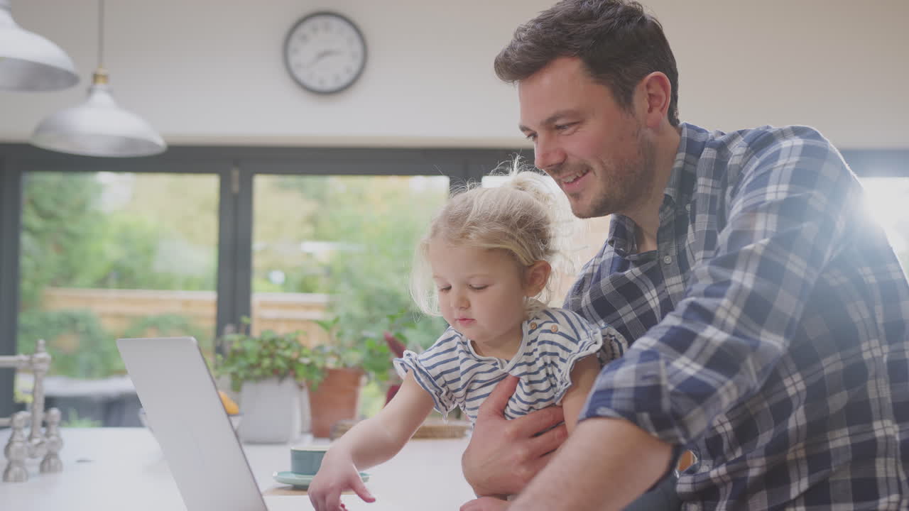 padre trabajando usando una computadora portátil en casa en el mostrador de la cocina mientras cuida a su hija pequeña - filmado en cámara lenta