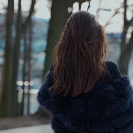 Attractive girl in the city park. Stylish woman in fur coat turning around and looking on camera outdoors. Slow motion.