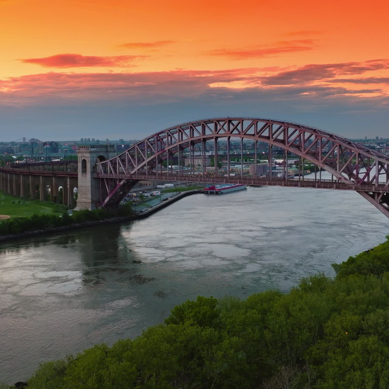 Hell Gate Bridge connecting two green banks. Picturesque structure at the backdrop of orange sky at sun dusk