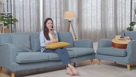 full body of asian teen girl recording voice message on smartphone while sitting on sofa in the living room at home