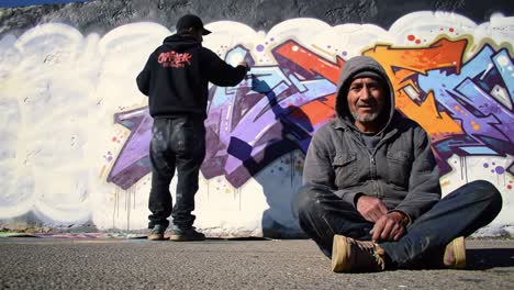 two artists engage in a creative process within a city park. one is painting a large graffiti mural against a colorful wall, while the other sits nearby, observing the artistic display.