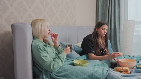 two young girls sit in bed covered with teal blanket, enjoying breakfast together, one holds mug and toast while other touches her face, assorted snacks, and drinks are placed on blanket in front