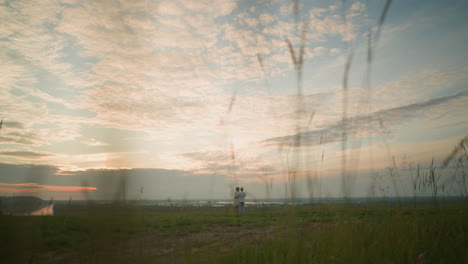 a couple stands on a grassy hill beside a tranquil lake at sunset, gazing at the sky. the man, in a white shirt, hat, and jeans, holds the woman by the waist. she wears a black hat and a white dress