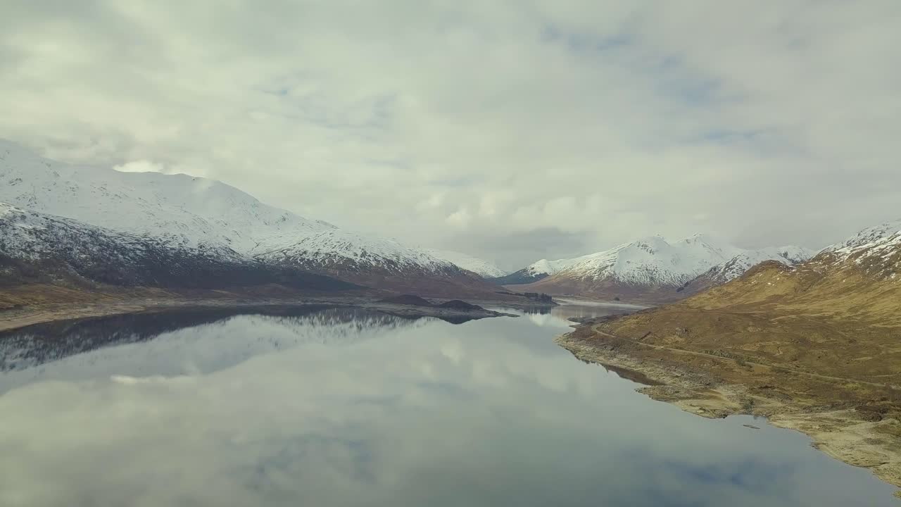 Aerial panorama view showing beautiful lake in Scottish highlands with snowy mountains in background and dense clouds at sky