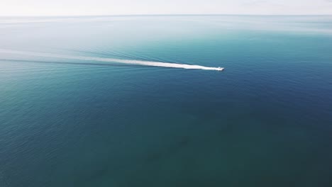 Drone-clip-of-wide-open-blue-sea-with-white-boat-moving-from-left-to-right-leaving-rippling-boat-wake