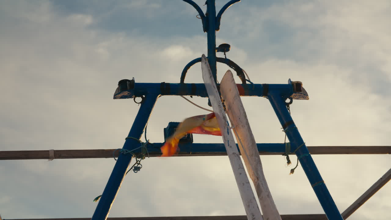 The Spanish flag waves in the wind against a blue fishing boat frame during golden hour. Strong motion and coastal atmosphere