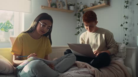 dos adolescentes caucásicos sentados en la cama y aprendiendo de libros y computadoras portátiles juntos