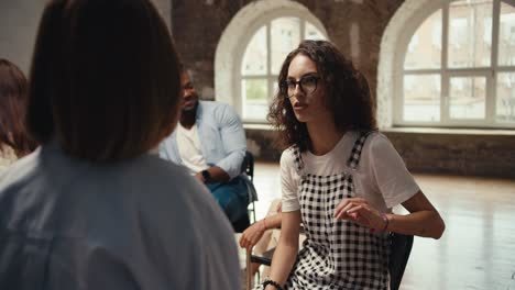 Ein-Brünettes-Mädchen-Mit-Lockigem-Haar,-Brille-Und-Kariertem-Overall-Kommuniziert-Paarweise-Während-Einer-Gruppentherapie-In-Einem-Braunen-Saal