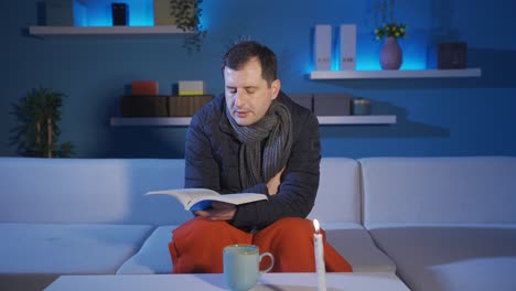 el hombre sentado en el sofá en casa está leyendo un libro a la luz de las velas, tiene frío en el frío, no hay gas, la casa no se calienta.