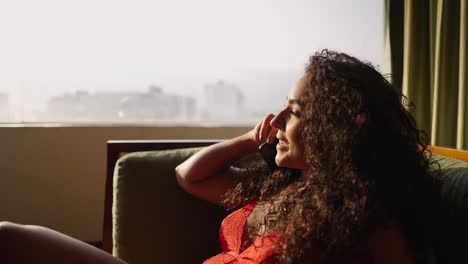 woman with curly hair talking on phone by a window