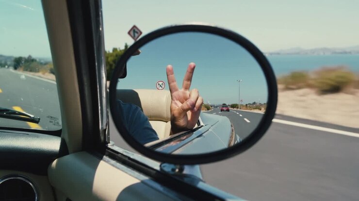 Auto che guida su un'autostrada costiera mentre l'avambraccio con camicia blu fa il segno della V nello specchio rotondo, passando il mare.