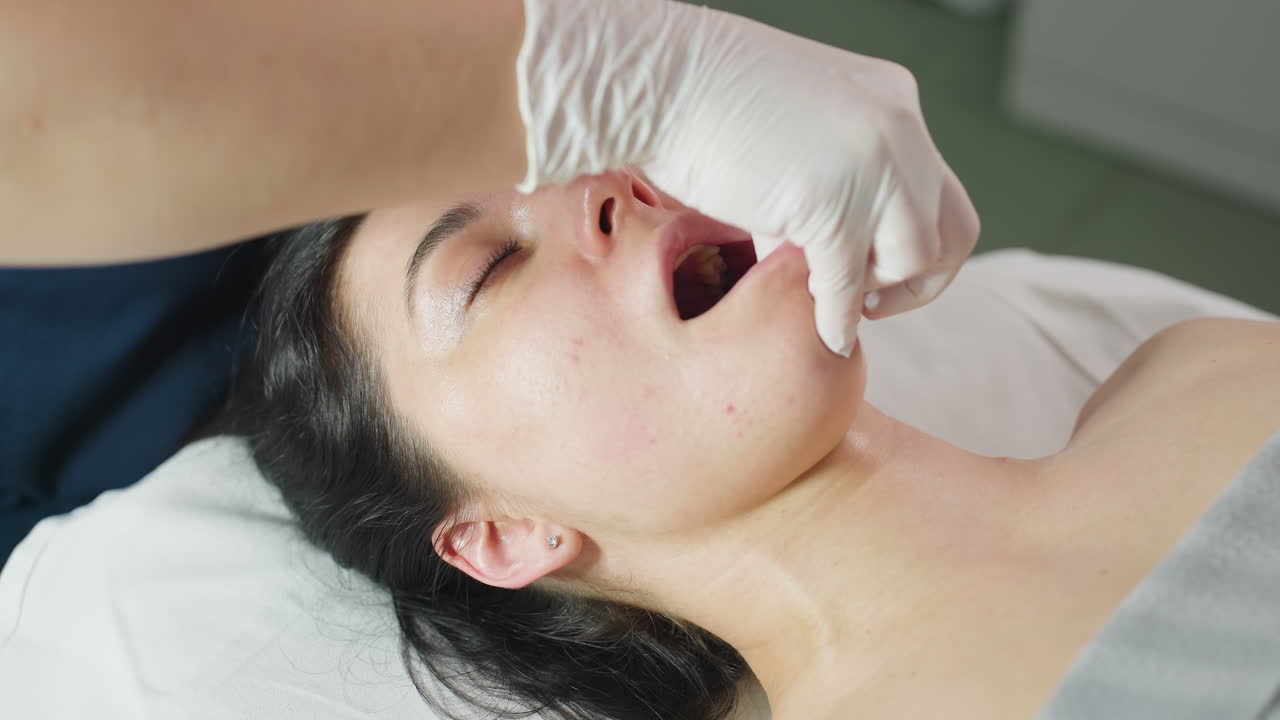 Close up of female visitor lying on massage chair opens mouth and closes eyes gently, tenses slightly then releases as expert wearing protective gloves dips fingertips into mouth massaging lips