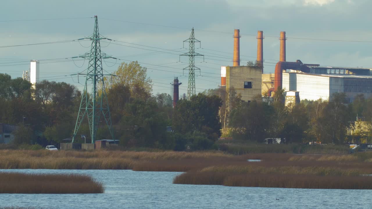 вид издалека на заброшенный завод по выплавке металла, красные кирпичные трубы и линии электропередач в солнечный осенний день, широкий снимок с расстояния над озером лиепая
