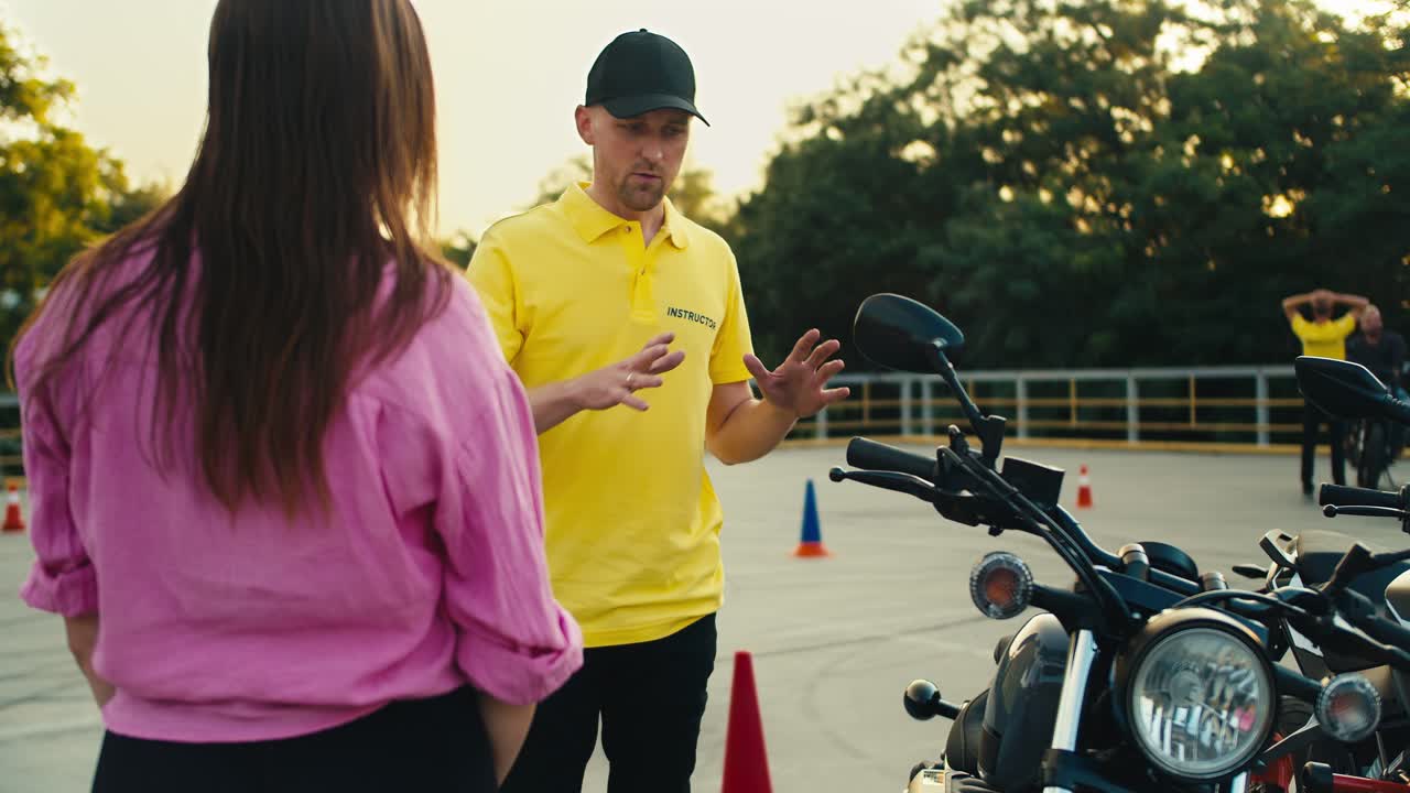 A driving instructor in a yellow t-shirt tells a female student in a pink t-shirt how to behave while driving a motorcycle