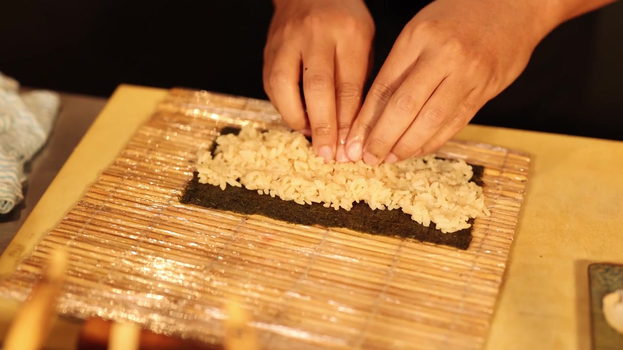 Primo piano di mani che dispongono con cura riso condito sull'alga nori su una stuoia di bambù a Bangkok.