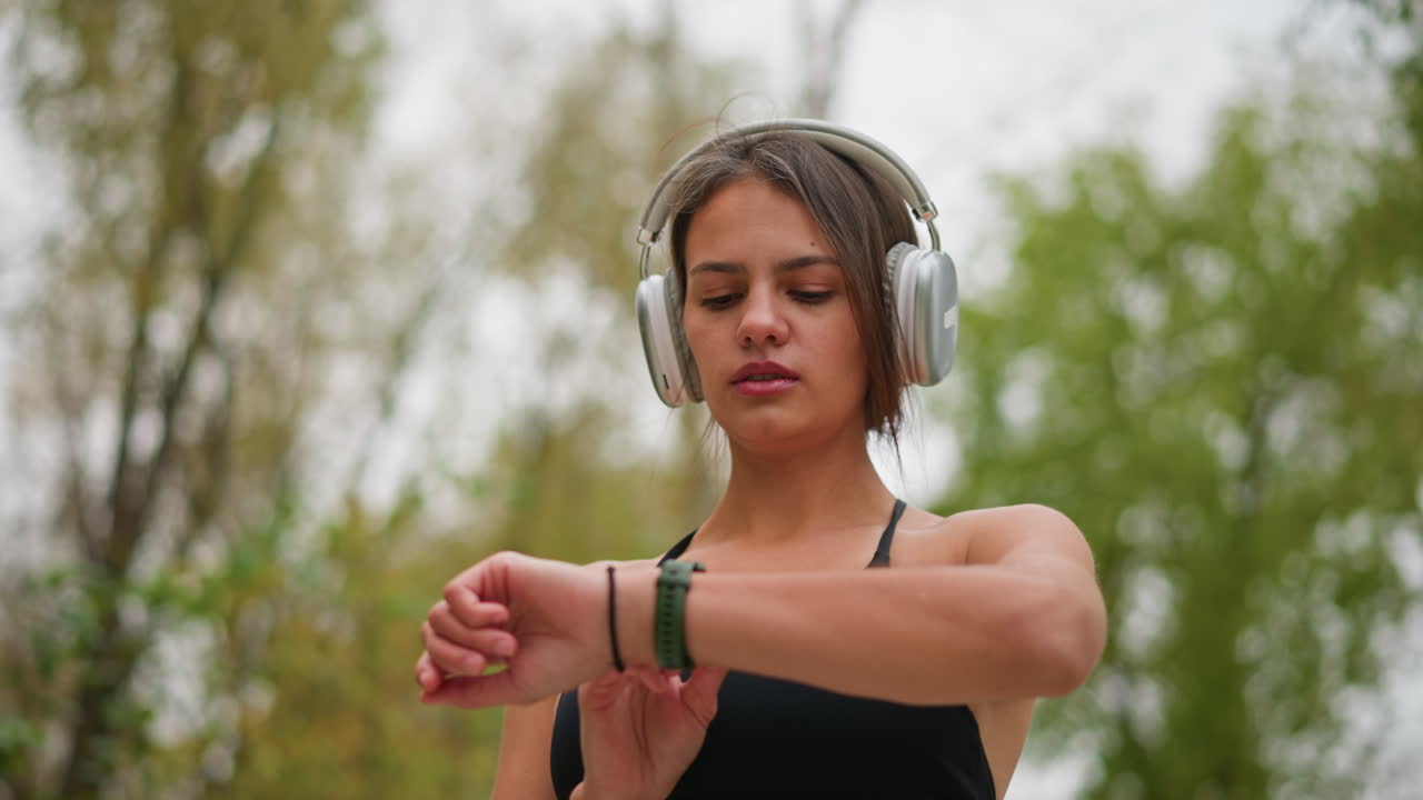 Ritratto di una giovane donna che indossa le cuffie, concentrata sul suo orologio da polso verde, profondamente impegnata a controllare l'ora durante una sessione di fitness all'aperto, che riflette consapevolezza e gestione del tempo.