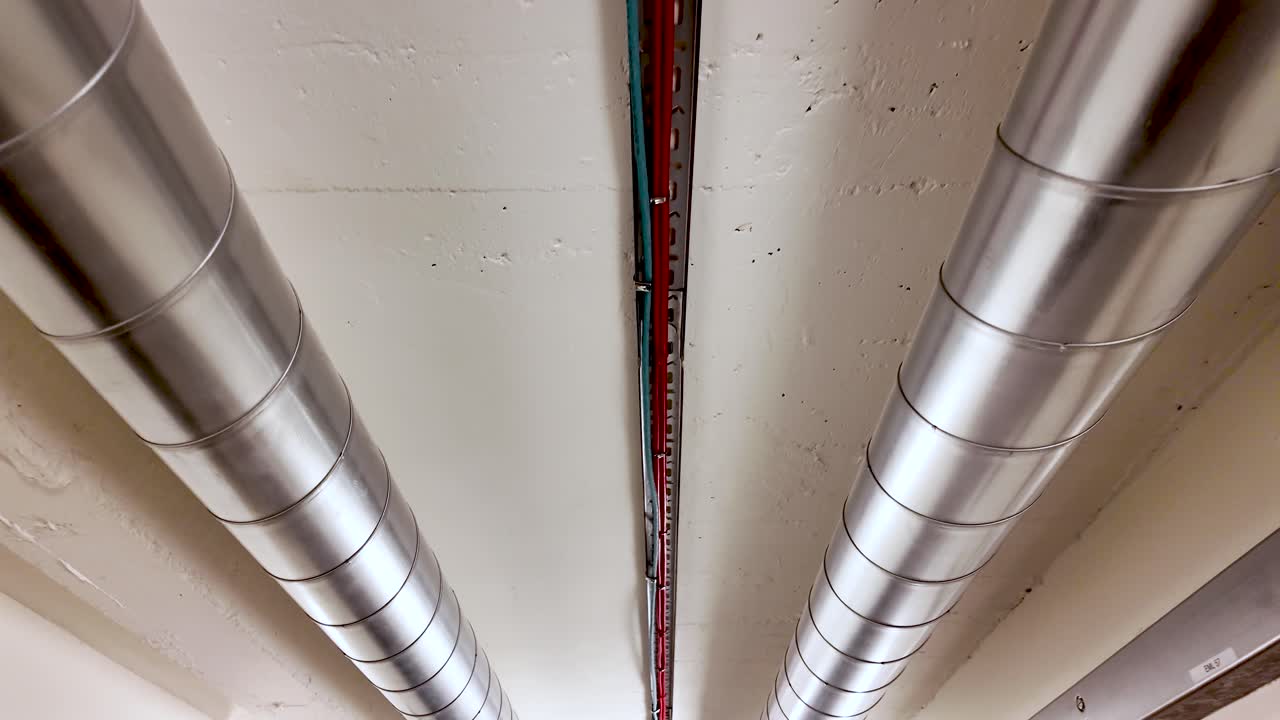 Coppia di tubi metallici di canalizzazione e cablaggio che attraversano un sistema di ventilazione industriale con soffitto bianco, aria condizionata e installazione elettrica